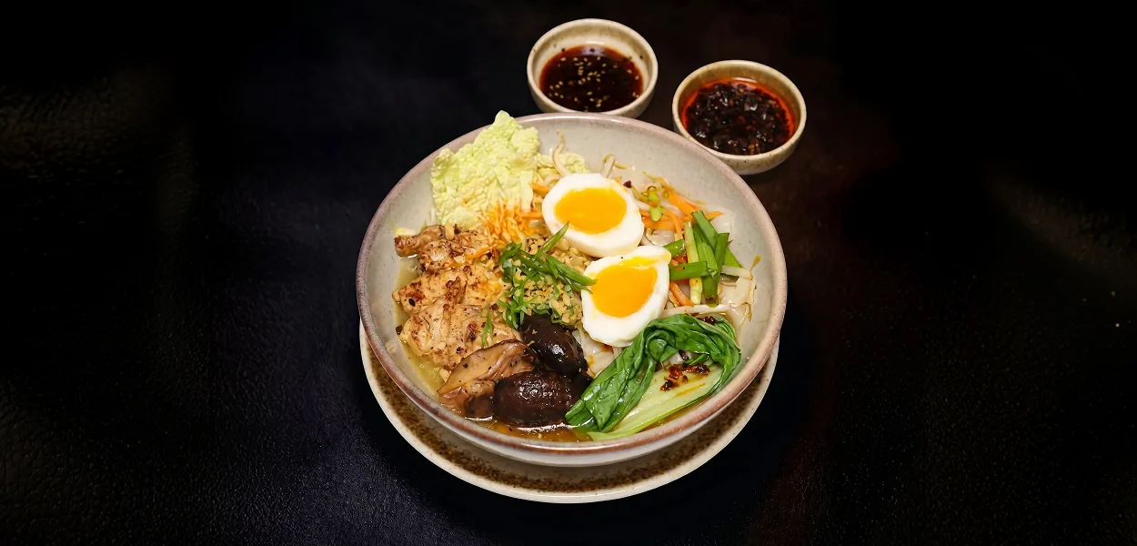 Chicken noodle bowl with soft-boiled eggs, mushrooms and pak choi beside two chilli sauces at Nara Thai