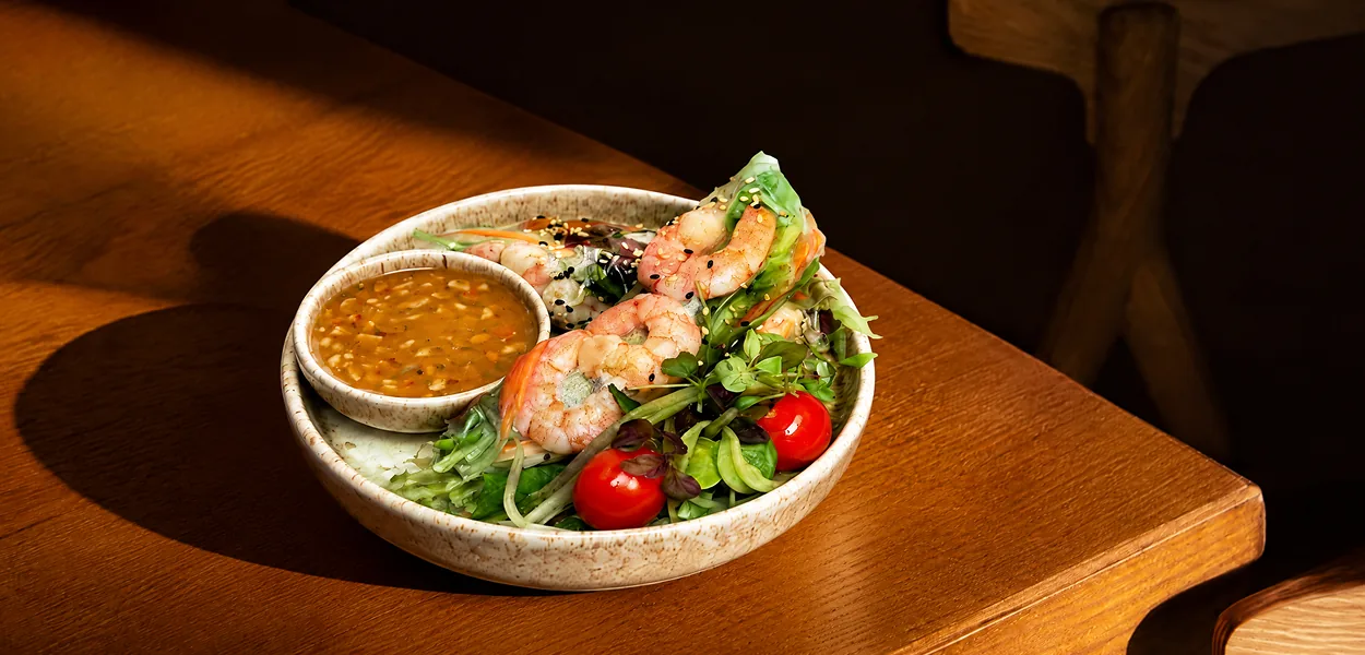 Prawn summer rolls with salad leaves, cherry tomatoes and a bowl of dipping sauce on a wooden table at Nara Thai