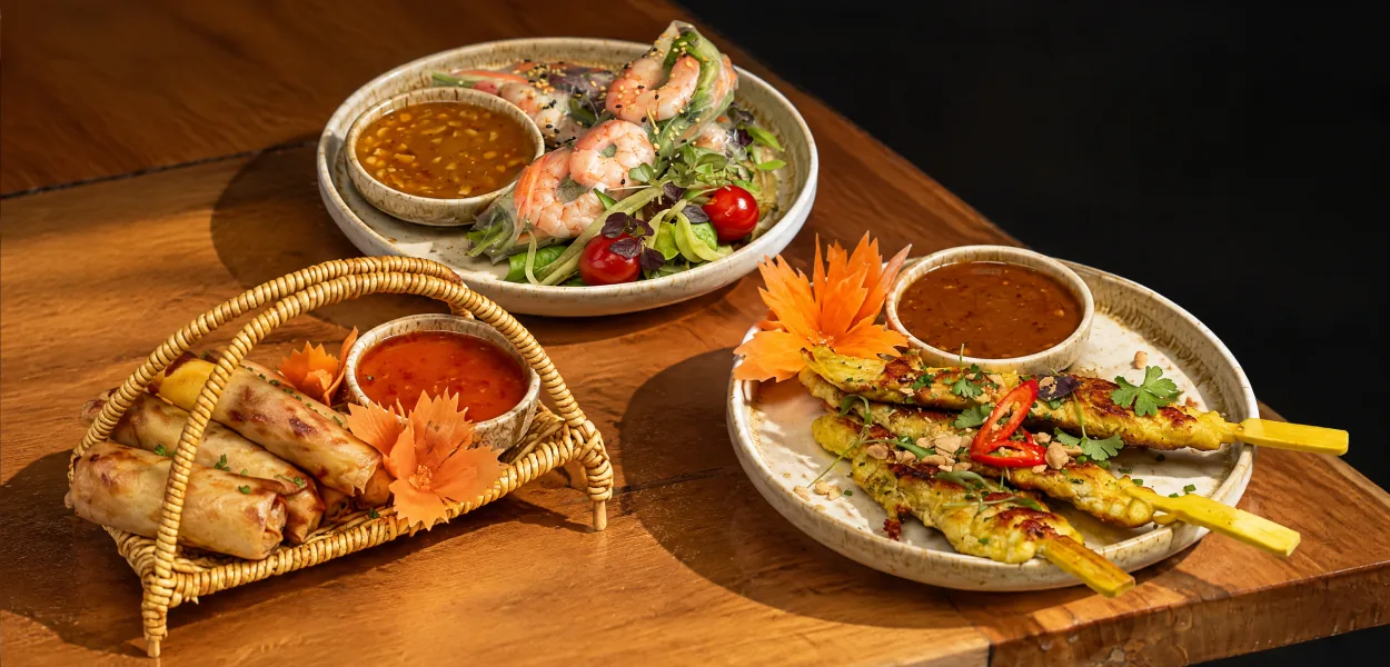 Spring rolls, prawn summer rolls and grilled satay skewers with dipping sauces on a wooden table at Nara Thai