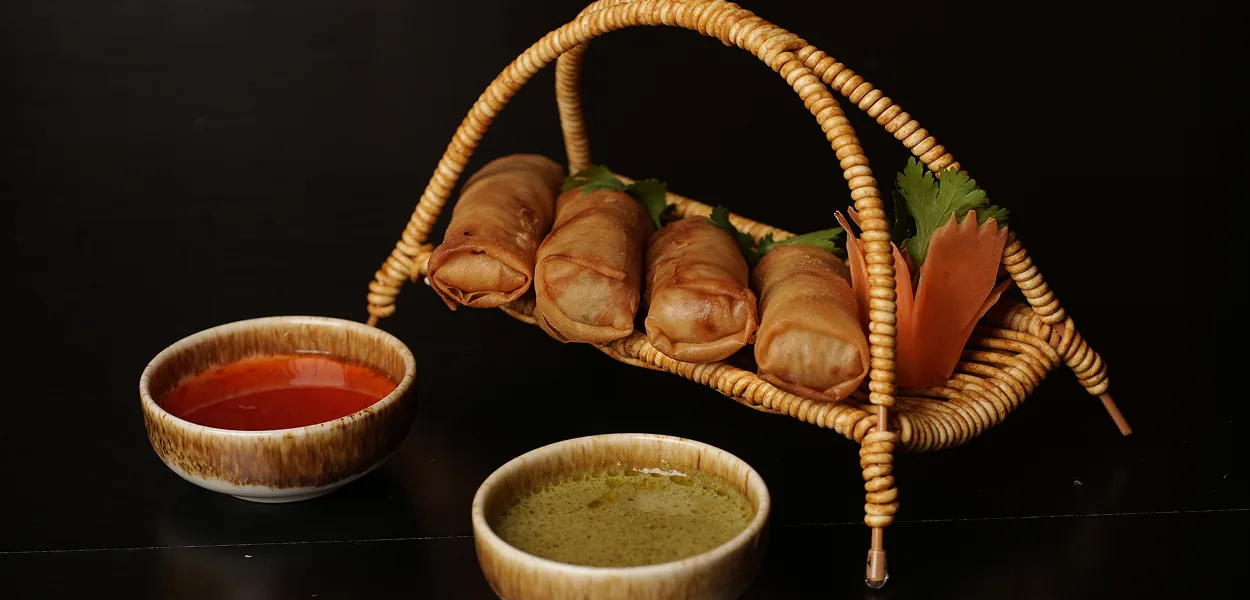 Four fried spring rolls in a woven basket stand with red and green dipping sauces at Nara Thai