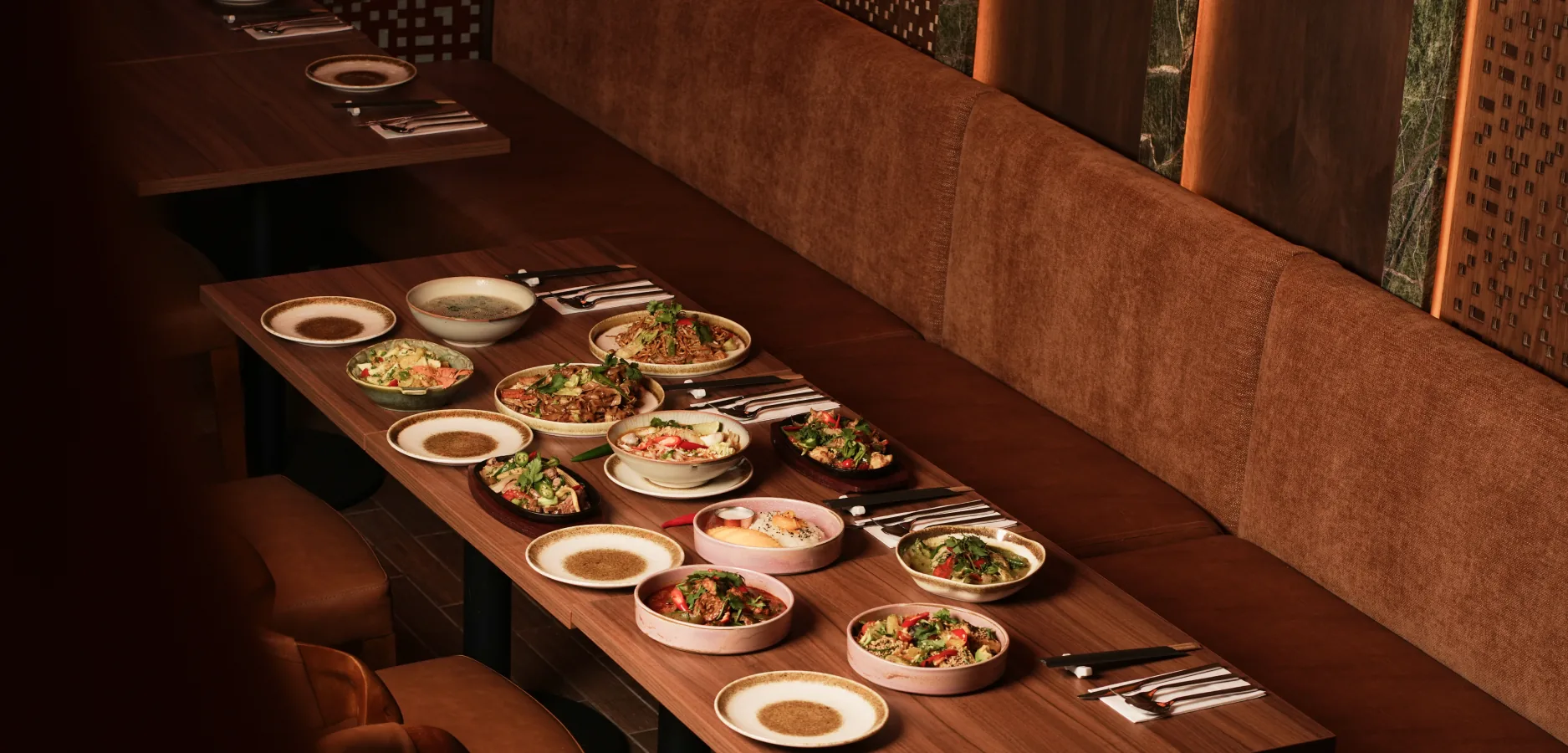 Long wooden table beside a banquette laid with Thai noodle dishes, curries, a soup and sizzling plates at Nara Thai